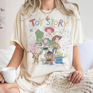 Puede incluir: Camiseta color crema con las palabras "Toy Story" en letras coloridas, junto con personajes de dibujos animados como Woody, Buzz Lightyear y Rex. El dise&ntilde;o incluye garabatos divertidos y la frase "yee haw".