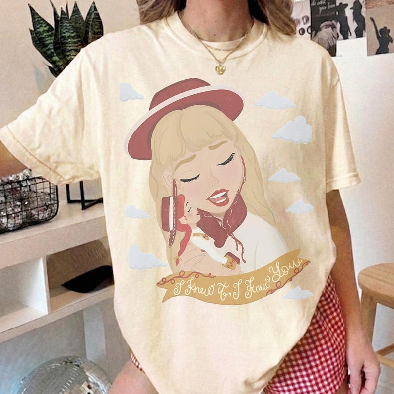 May include: A cream-colored short-sleeve t-shirt featuring a graphic illustration of a blonde character wearing a red hat and holding a small doll. The design includes scattered white clouds and a banner with the text I Knew It, I Knew You.