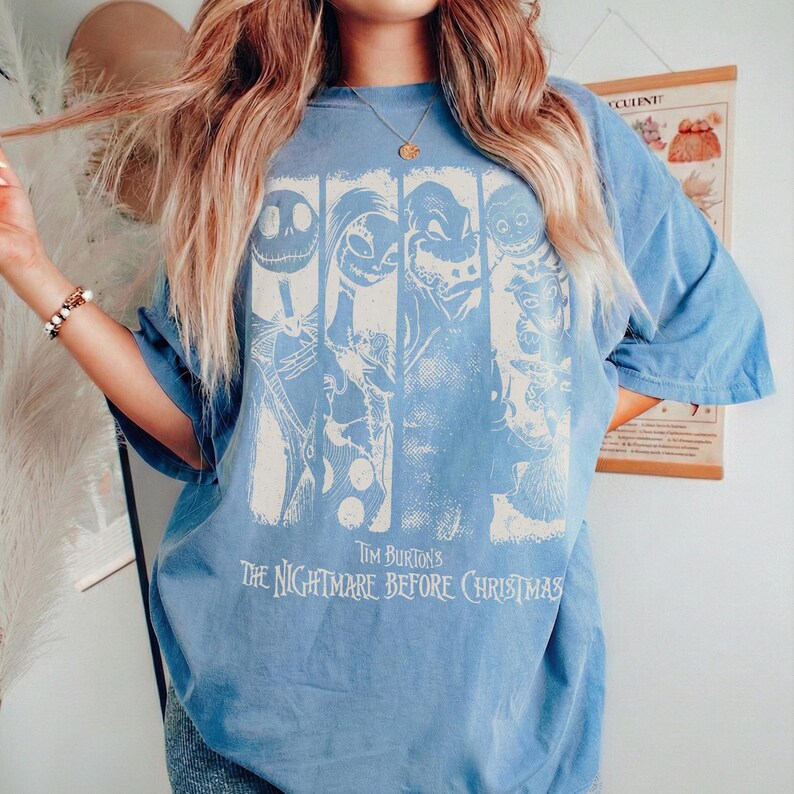 May include: A blue short-sleeve t-shirt featuring a white graphic print of characters from The Nightmare Before Christmas. The design includes four vertical panels with character illustrations and the text Tim Burton's The Nightmare Before Christmas.