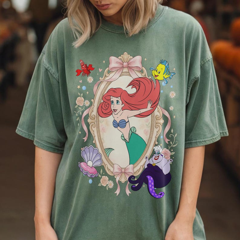 Puede incluir: Camiseta verde con una ilustraci&oacute;n enmarcada de Ariel de La Sirenita, con Flounder, Sebasti&aacute;n y &Uacute;rsula. La ilustraci&oacute;n incluye un lazo rosa, flores y una concha. La camiseta es de color verde apagado.
