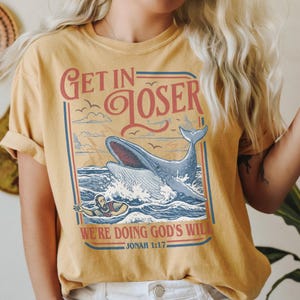 Puede incluir: Camiseta de color amarillo mostaza con un dise&ntilde;o gr&aacute;fico que presenta una ballena rompiendo la superficie del oc&eacute;ano. El texto "Get in Loser" est&aacute; encima de la ballena, y "We're doing God's will" est&aacute; debajo. La camiseta tambi&eacute;n incluye el vers&iacute;culo b&iacute;blico Jon&aacute;s 1:17.