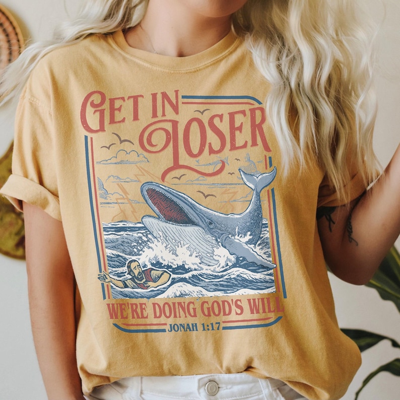 Puede incluir: Camiseta de color amarillo mostaza con un dise&ntilde;o gr&aacute;fico que presenta una ballena rompiendo la superficie del oc&eacute;ano. El texto "Get in Loser" est&aacute; encima de la ballena, y "We're doing God's will" est&aacute; debajo. La camiseta tambi&eacute;n incluye el vers&iacute;culo b&iacute;blico Jon&aacute;s 1:17.