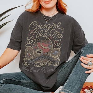 May include: A charcoal gray short-sleeve t-shirt featuring a graphic print with the text Cowgirl Like Me, Eyes Full of Stars, and Takes One to Know One. The design includes a cowboy hat, a horseshoe, a rope border, and musical notes.