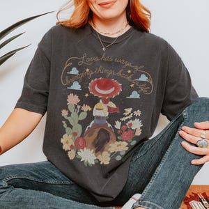 May include: A charcoal gray t-shirt featuring a graphic of a cowgirl figure wearing a red hat, sitting on a brown horse. The design includes cacti, flowers, clouds, stars, and the text "Love has ways of bringing things back to life" in script.