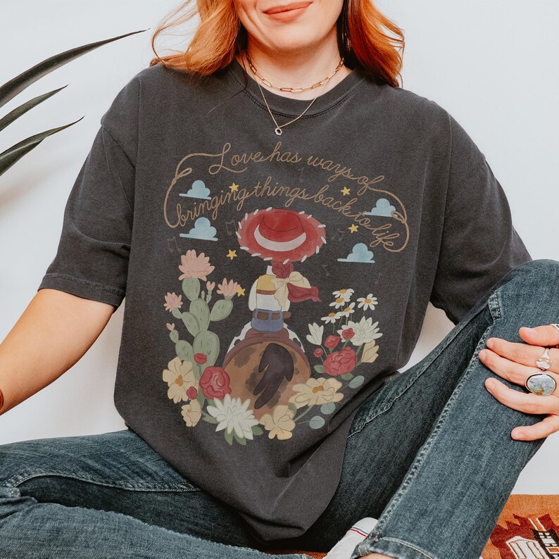 May include: A charcoal gray t-shirt featuring a graphic of a cowgirl figure wearing a red hat, sitting on a brown horse. The design includes cacti, flowers, clouds, stars, and the text "Love has ways of bringing things back to life" in script.