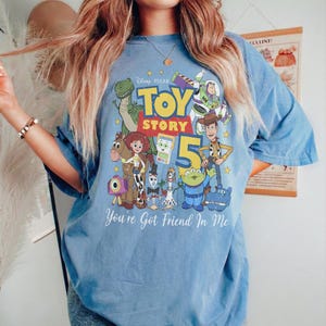 May include: A blue short-sleeve t-shirt featuring a colorful graphic print of Toy Story characters including Woody, Buzz Lightyear, Rex, and Jessie. The design includes the text Toy Story 5 and You've Got A Friend In Me in white lettering.