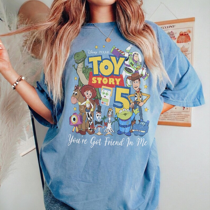 May include: A blue short-sleeve t-shirt featuring a colorful graphic print of Toy Story characters including Woody, Buzz Lightyear, Rex, and Jessie. The design includes the text Toy Story 5 and You've Got A Friend In Me in white lettering.