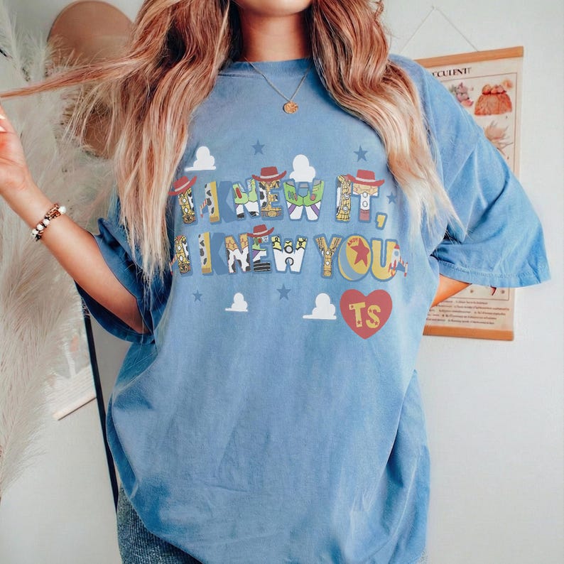 K&ouml;nnte beinhalten: Ein hellblaues Kurzarm-T-Shirt mit dem Text I KNEW IT, I KNEW YOU in einer mehrfarbigen Schriftart mit charakterbezogenen Mustern. Das Design enth&auml;lt kleine wei&szlig;e Wolkensymbole, blaue Sterne und ein rotes Herz mit den Buchstaben TS darin.