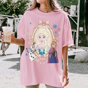 May include: Pink t-shirt featuring a framed portrait of Elsa from Frozen, with Anna and Olaf. The design includes a pink bow, snowflakes, and floral accents. The shirt is a light pink color.