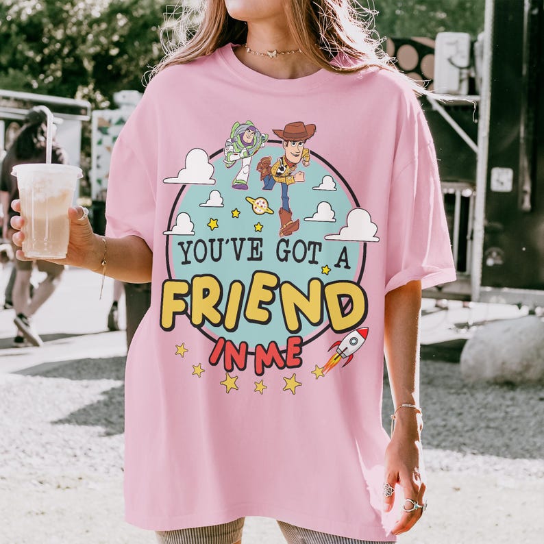 Puede incluir: Una camiseta rosa claro con un gr&aacute;fico circular de Buzz Lightyear y Woody de Toy Story. El dise&ntilde;o incluye nubes blancas, estrellas amarillas, un peque&ntilde;o cohete y el texto YOU'VE GOT A FRIEND IN ME en letras grandes amarillas y rojas.