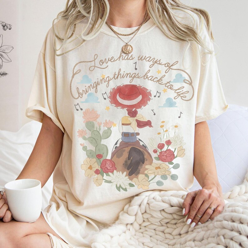 May include: A cream-colored short-sleeve t-shirt featuring a graphic print of a cowgirl on a horse surrounded by cacti and flowers. The text reads, Love has ways of bringing things back to life, accompanied by musical notes and small blue clouds.