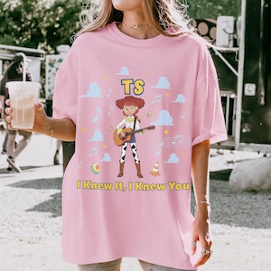 Puede incluir: Una camiseta rosa claro con el gr&aacute;fico de una vaquera de dibujos animados tocando una guitarra ac&uacute;stica. El dise&ntilde;o incluye nubes azules, notas musicales, una pelota, un cono de tr&aacute;fico y el texto TS e I Knew It, I Knew You en amarillo.