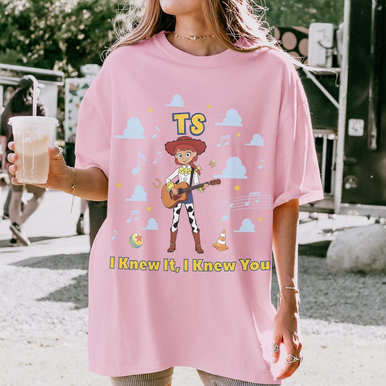 Puede incluir: Una camiseta rosa claro con el gr&aacute;fico de una vaquera de dibujos animados tocando una guitarra ac&uacute;stica. El dise&ntilde;o incluye nubes azules, notas musicales, una pelota, un cono de tr&aacute;fico y el texto TS e I Knew It, I Knew You en amarillo.