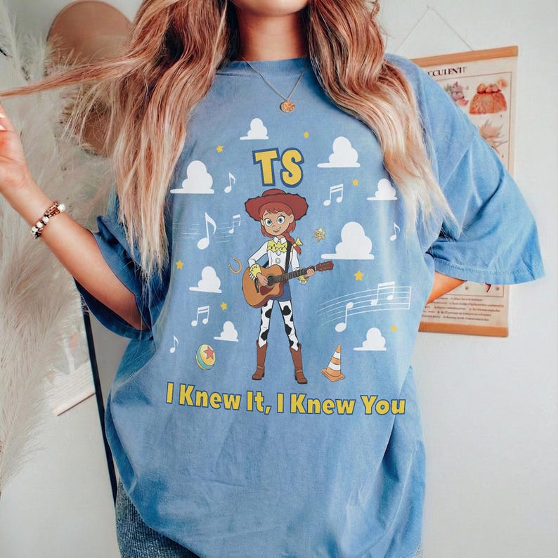 Puede incluir: Una camiseta azul claro con un gr&aacute;fico de una vaquera tocando una guitarra ac&uacute;stica. El dise&ntilde;o incluye el texto TS y I Knew It, I Knew You, rodeado de nubes blancas, notas musicales, una pelota amarilla y un cono de tr&aacute;fico.