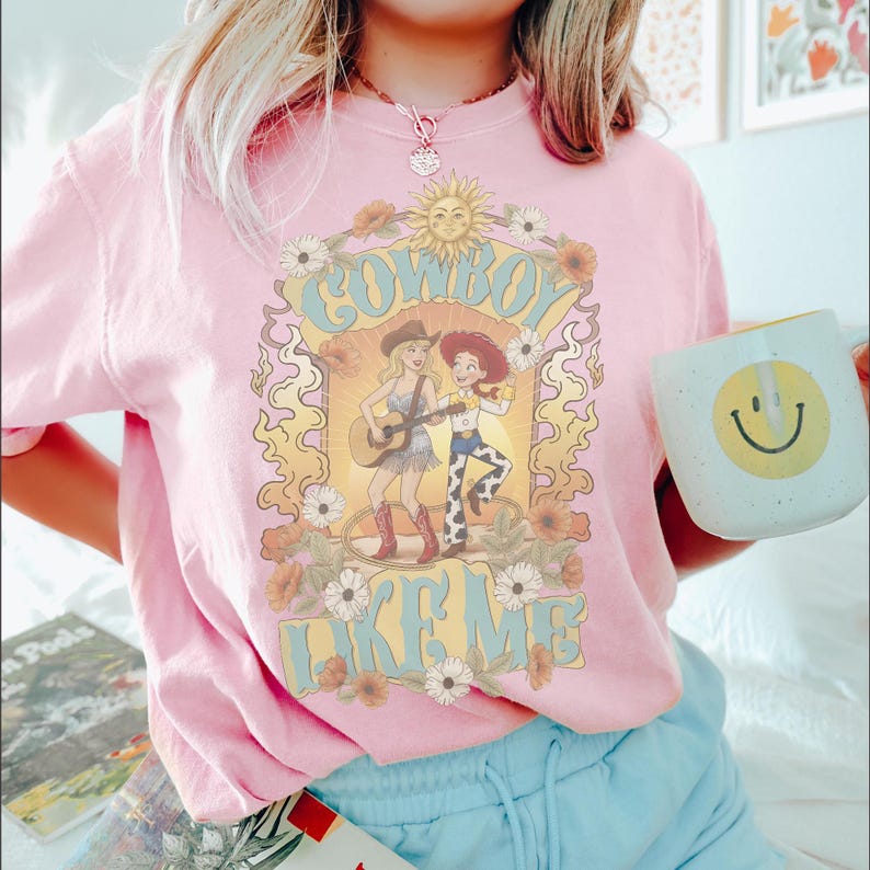 May include: A light pink t-shirt featuring a graphic print of two figures in cowboy attire, one playing a guitar, surrounded by a sun, flowers, and swirling flame-like patterns. The text "COWBOY LIKE ME" is printed in large, light blue letters.