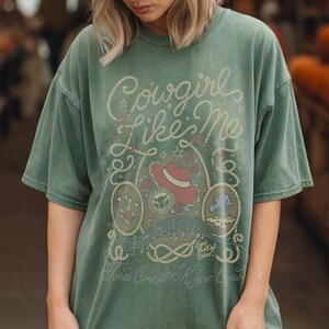 May include: A sage green short-sleeve t-shirt featuring a graphic print with the text Cowgirl Like Me, Eyes full of stars, and Takes One to Know One. The design includes illustrations of a red cowboy hat, a horseshoe, and rope motifs.