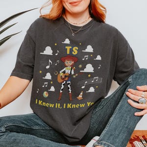 Puede incluir: Una camiseta gris carb&oacute;n de manga corta con un gr&aacute;fico de una vaquera tocando una guitarra ac&uacute;stica. El dise&ntilde;o incluye el texto TS y I Knew It, I Knew You, rodeado de nubes blancas, notas musicales, estrellas, una pelota y un cono de tr&aacute;fico.