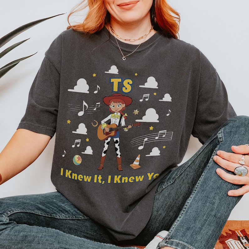 Puede incluir: Una camiseta gris carb&oacute;n de manga corta con un gr&aacute;fico de una vaquera tocando una guitarra ac&uacute;stica. El dise&ntilde;o incluye el texto TS y I Knew It, I Knew You, rodeado de nubes blancas, notas musicales, estrellas, una pelota y un cono de tr&aacute;fico.