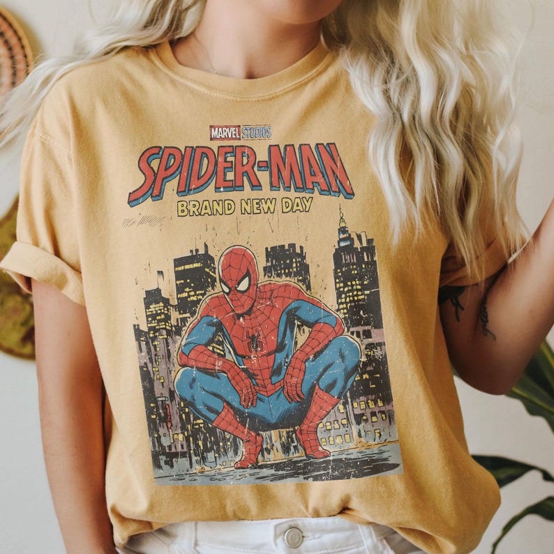 May include: Mustard yellow t-shirt featuring a vintage Spider-Man comic book graphic. The design includes the words "Marvel Studios Spider-Man Brand New Day" with an illustration of Spider-Man crouching in front of a city skyline.