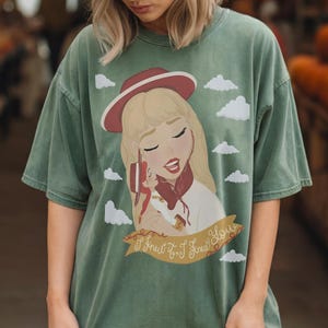 May include: A sage green t-shirt featuring a graphic illustration of a blonde character wearing a red hat, holding a small doll, surrounded by white clouds. A tan banner at the bottom displays the cursive text, I Knew It, I Knew You.