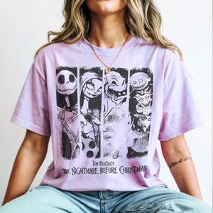 May include: A light purple short-sleeve t-shirt featuring a black graphic print of characters from Tim Burton's The Nightmare Before Christmas. The design includes four vertical panels showing Jack Skellington, Sally, Oogie Boogie, and Lock, Shock, and Barrel.