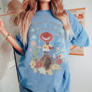 May include: A blue short-sleeve t-shirt featuring a graphic of a cowgirl on a horse surrounded by cacti and flowers. The text "Love has ways of bringing things back to life" is printed in gold script above the illustration with musical notes.