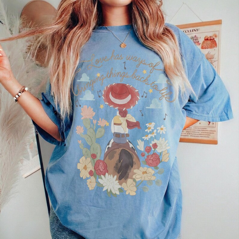 May include: A blue short-sleeve t-shirt featuring a graphic of a cowgirl on a horse surrounded by cacti and flowers. The text "Love has ways of bringing things back to life" is printed in gold script above the illustration with musical notes.