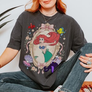 Puede incluir: Camiseta gris oscuro con una ilustraci&oacute;n enmarcada de Ariel de La Sirenita, con Flounder, Sebasti&aacute;n y &Uacute;rsula. El dise&ntilde;o incluye un lazo rosa, flores y una concha. La camiseta es una prenda informal.