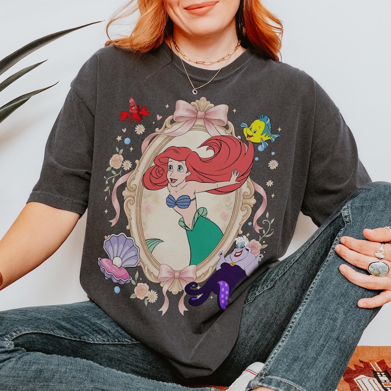 Puede incluir: Camiseta gris oscuro con una ilustraci&oacute;n enmarcada de Ariel de La Sirenita, con Flounder, Sebasti&aacute;n y &Uacute;rsula. El dise&ntilde;o incluye un lazo rosa, flores y una concha. La camiseta es una prenda informal.