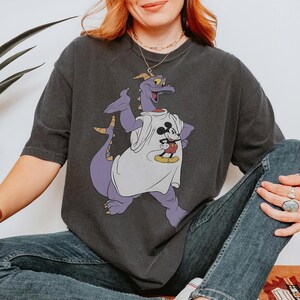 Peut inclure: T-shirt gris fonc&eacute; avec un dragon violet portant un t-shirt blanc avec un motif Mickey Mouse. Le dragon a des cornes jaunes et une longue queue. Le t-shirt est port&eacute; avec un jean bleu.