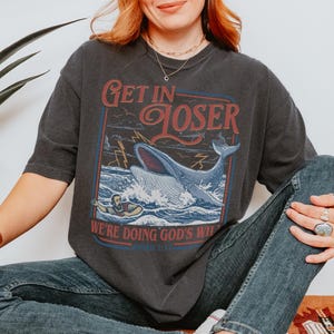 Puede incluir: Camiseta gris oscuro con un gr&aacute;fico que presenta una ballena rompiendo el oc&eacute;ano, rayos y el texto "Get in Loser, We're Doing God's Will". El dise&ntilde;o est&aacute; enmarcado en rojo y azul.