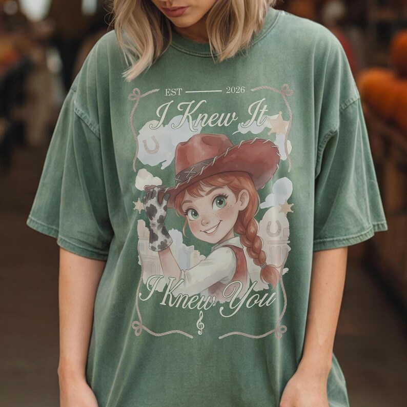 May include: A sage green short-sleeve t-shirt featuring a graphic print of a cartoon character wearing a brown cowboy hat and a cow-print glove. The text reads EST 2026, I Knew It, and I Knew You, surrounded by stars, clouds, and a treble clef.