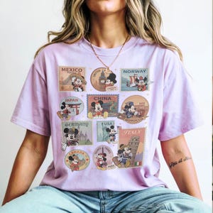 May include: A light purple t-shirt featuring postage stamp designs with Mickey Mouse in various countries, including Mexico, China, and Italy. The stamps are in a variety of colors, with the text of each country's name and Mickey Mouse's image.