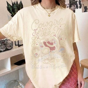 May include: A cream-colored short-sleeve t-shirt featuring a graphic print with the text "Cowgirl Like Me" and "Takes One to Know One." The design includes illustrations of a cowboy hat, roses, musical notes, and rope motifs in muted tones.