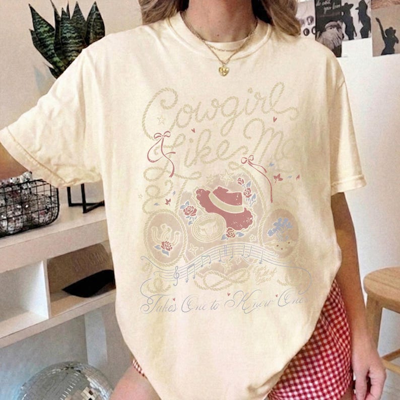 May include: A cream-colored short-sleeve t-shirt featuring a graphic print with the text "Cowgirl Like Me" and "Takes One to Know One." The design includes illustrations of a cowboy hat, roses, musical notes, and rope motifs in muted tones.