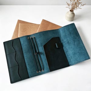 Peut inclure: Journal en cuir bleu sarcelle ouvert, avec une poche en cuir noir et une fermeture élastique. Le journal comporte plusieurs poches et une fermeture à cordon. Des cahiers en papier brun sont visibles en arrière-plan.