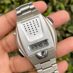 Op de afbeelding: Een vintage Seiko digitale horloge met een zilverkleurige kast en band. De wijzerplaat geeft de tijd weer als 9:40 uur. Het horloge heeft een reeks kleine gaatjes en twee zwarte knoppen. De band is gemaakt van metalen schakels.
