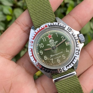 May include: Vintage military wristwatch with a green face, a red star, and a parachute emblem. The watch has a silver bezel with black and red markings and a green nylon strap. The watch face displays the numbers 1 to 12.