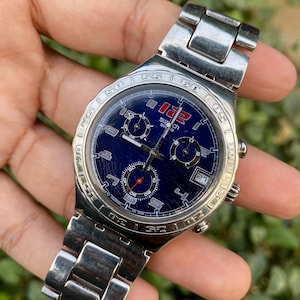 May include: A silver-toned Swatch watch with a blue face and a stainless steel band. The watch face features multiple sub-dials, a date display, and the Swatch logo. The watch is a chronograph style.