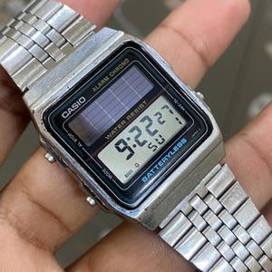 May include: A silver Casio digital watch with a rectangular face displaying the time 9:22:27. The watch has a stainless steel band and features the text "CASIO", "ALARM CHRONO", and "WATER RESIST". The watch is battery-less.