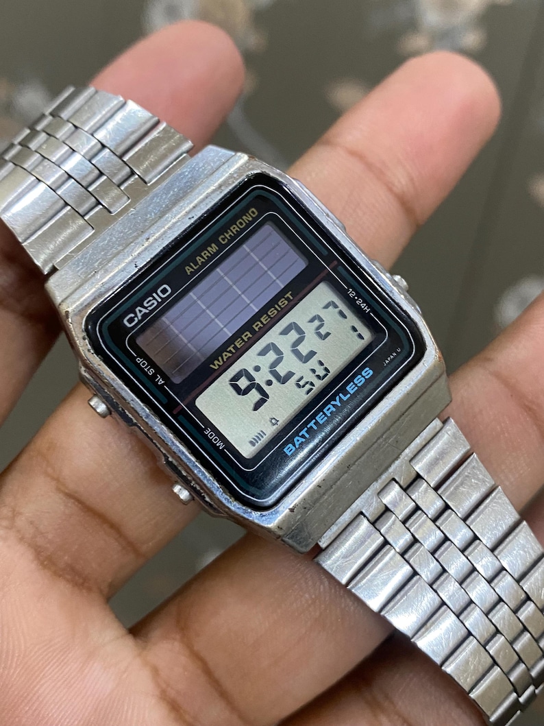 May include: A silver Casio digital watch with a rectangular face displaying the time 9:22:27. The watch has a stainless steel band and features the text "CASIO", "ALARM CHRONO", and "WATER RESIST". The watch is battery-less.