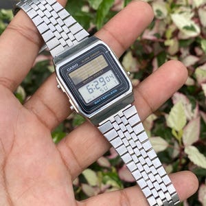 Może przedstawiać: Srebrny cyfrowy zegarek Casio z prostokątną czarną tarczą wyświetlającą godzinę 6:29:04. Zegarek ma srebrną metalową bransoletę, a na tarczy widnieją napisy "CASIO" i "BATTERYLESS". Zegarek jest wodoodporny.