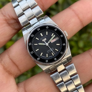 May include: A silver-toned Seiko 5 automatic wristwatch with a black dial. The watch features a day and date display, with the day reading "SUN 13". The watch band is made of silver-toned metal links.
