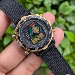 Puede incluir: Un reloj digital Citizen con una correa de nailon negra. La esfera del reloj es negra con detalles dorados y muestra la hora. El reloj tiene múltiples funciones, incluyendo un cronómetro y un temporizador. El reloj es resistente al agua hasta 100 metros.