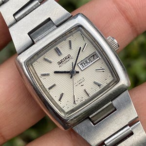 Puede incluir: Un reloj de pulsera automático Seiko vintage con esfera plateada y correa de acero inoxidable. La esfera muestra el día y la fecha, con el día que indica "JUE 7". El reloj tiene una caja rectangular y una esfera texturizada.