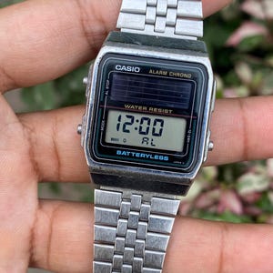 May include: Silver-toned Casio digital watch with a rectangular face. The watch displays the time as 12:00 and features the text "WATER RESIST" and "BATTERYLESS". The watch has a silver link band.