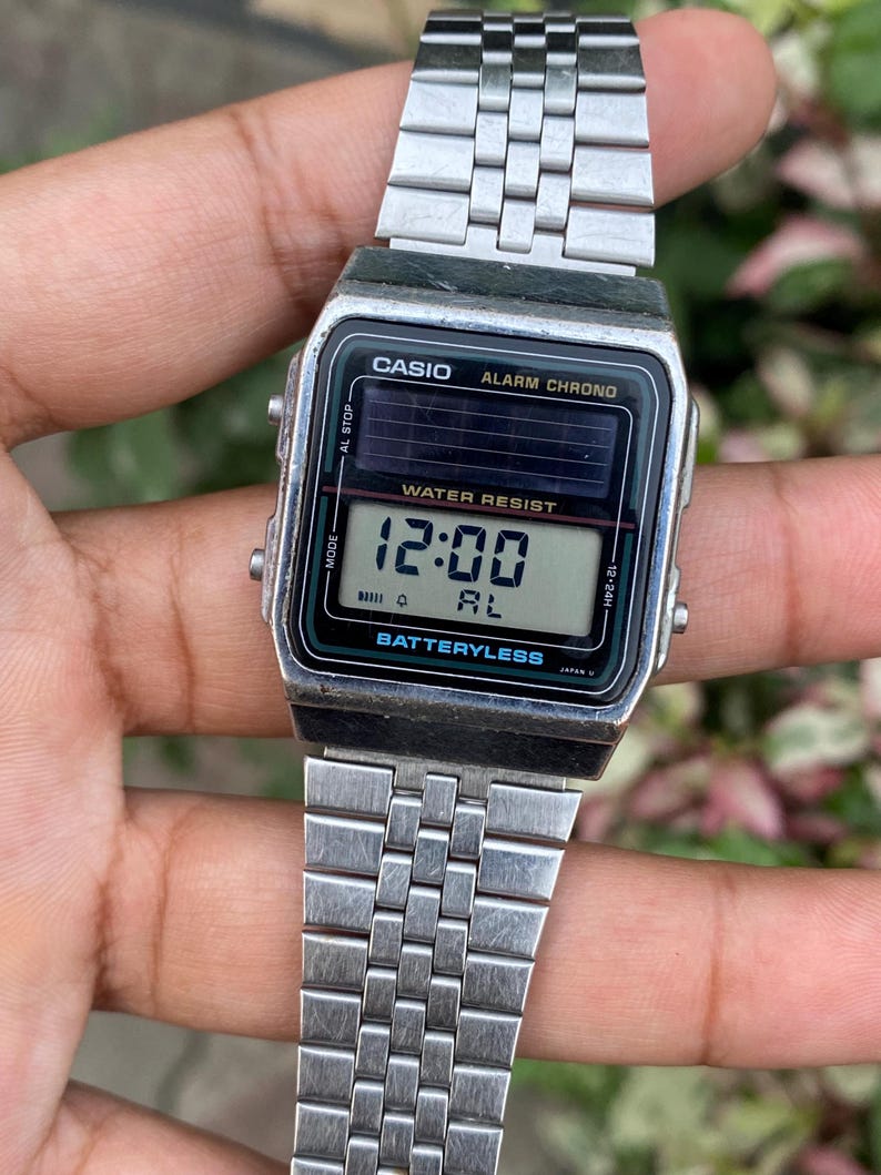 May include: Silver-toned Casio digital watch with a rectangular face. The watch displays the time as 12:00 and features the text "WATER RESIST" and "BATTERYLESS". The watch has a silver link band.