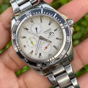 May include: A silver-tone Casio Quartz analog watch with a white textured dial, a black rotating bezel, and a stainless steel link bracelet. The watch face features three sub-dials, a dolphin logo, and the text CASIO QUARTZ WR 200M.