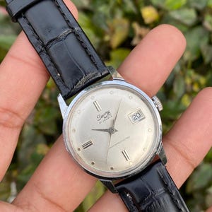Könnte beinhalten: Eine silberfarbene Vintage-Armbanduhr von Sacony mit Automatikwerk und einem schwarzen Lederarmband. Das Zifferblatt der Uhr ist silberfarben mit schwarzen Zeigern und Markierungen sowie einer Datumsanzeige auf 3 Uhr. Die Uhr ist mit "Shock Protected" beschriftet.