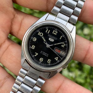 May include: A silver-tone stainless steel Seiko 5 automatic wristwatch with a black dial, featuring white Arabic numerals, luminous hands, a day-date window at the three o'clock position, and a metal link bracelet with a fold-over clasp.